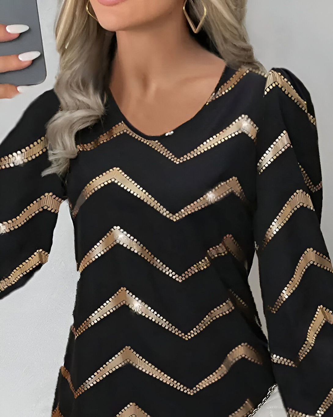 Une femme porte un chemisier noir à manches longues. Le chemisier est orné de sequins dorés. La femme a les cheveux blonds et les ongles blancs. Elle porte des boucles d'oreilles en forme de cœur.