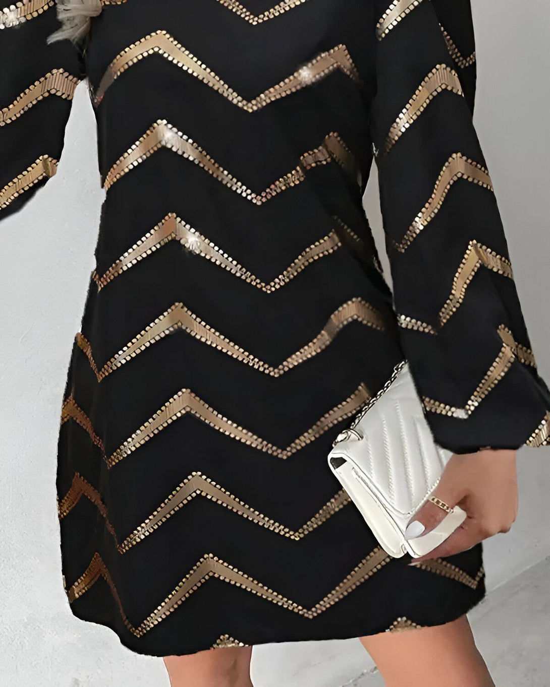 Une femme porte une robe noire à sequins dorés. La robe est courte et a des manches longues. La femme porte également un sac à main blanc.