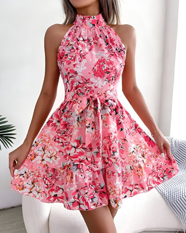 Une jeune femme porte une robe rose à fleurs. La robe est courte et évasée, avec un col montant et des bretelles fines. La femme est debout, les mains sur les hanches. Elle a les cheveux longs et blonds et porte un maquillage léger.