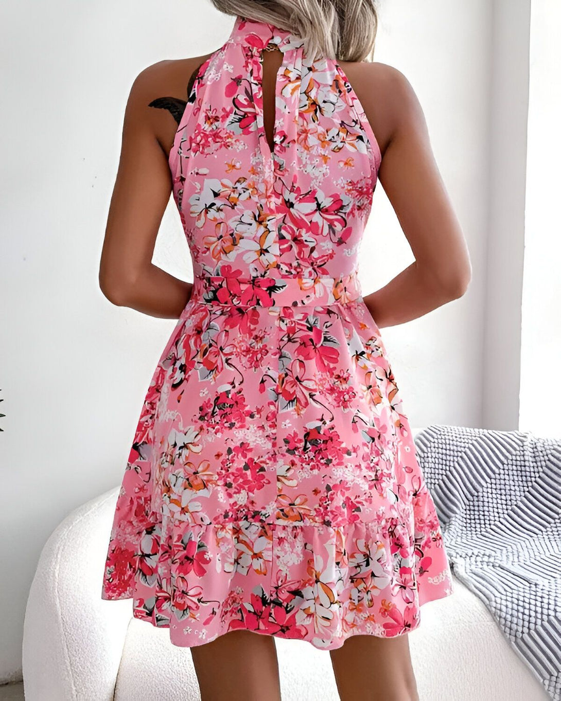 Une femme porte une robe rose sans manches à motifs floraux. La robe est courte et évasée, avec un col montant et une encolure en V dans le dos.