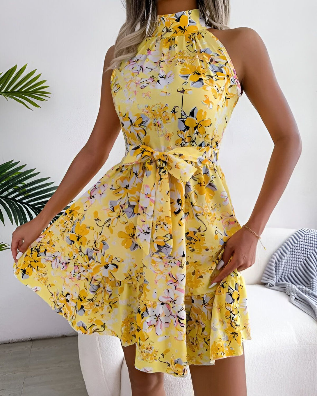 Une jeune femme porte une robe jaune à fleurs. La robe est courte et évasée, avec un col montant et des bretelles fines. La femme est debout, les cheveux lâchés et le sourire aux lèvres.