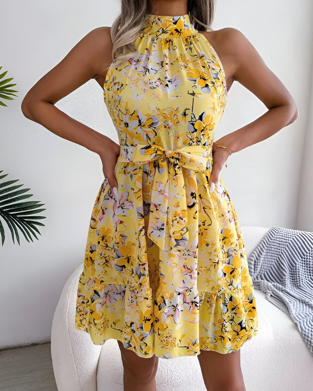 Une jeune femme porte une robe jaune à fleurs. La robe est courte et évasée, avec un col montant et des bretelles fines. La femme est debout, les mains sur les hanches. Elle a les cheveux longs et blonds, et elle est maquillée avec un maquillage naturel.