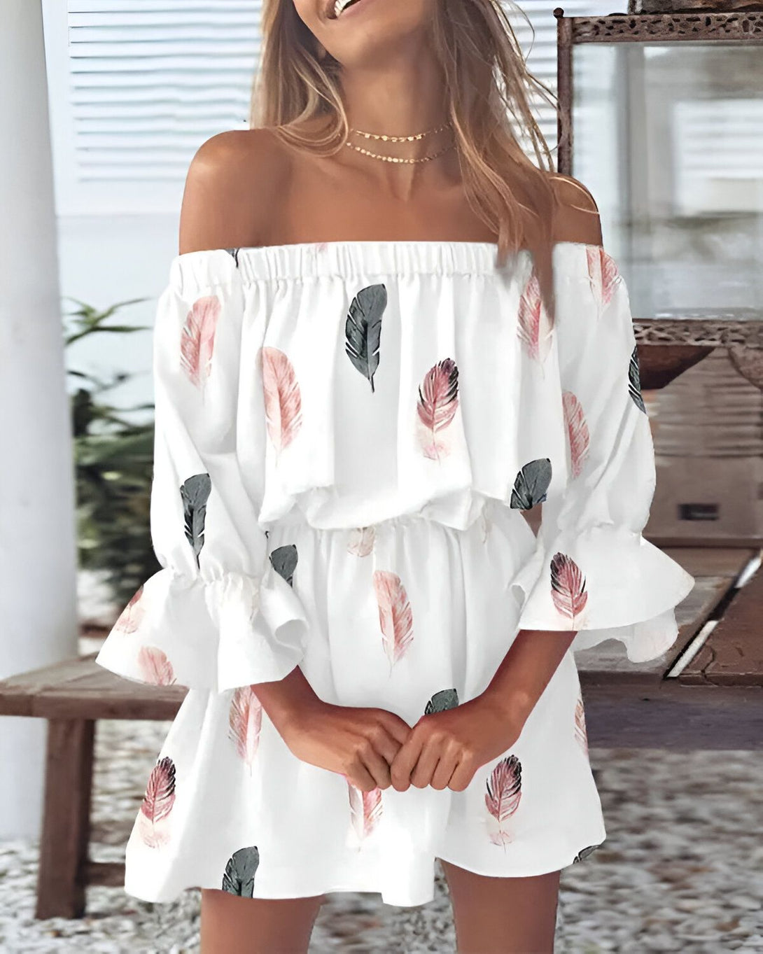 Une jeune femme porte une robe blanche à épaules dénudées. La robe est imprimée de plumes roses et vertes. La femme a les cheveux longs et blonds et elle porte un collier doré. Elle est debout dans un jardin et elle sourit.