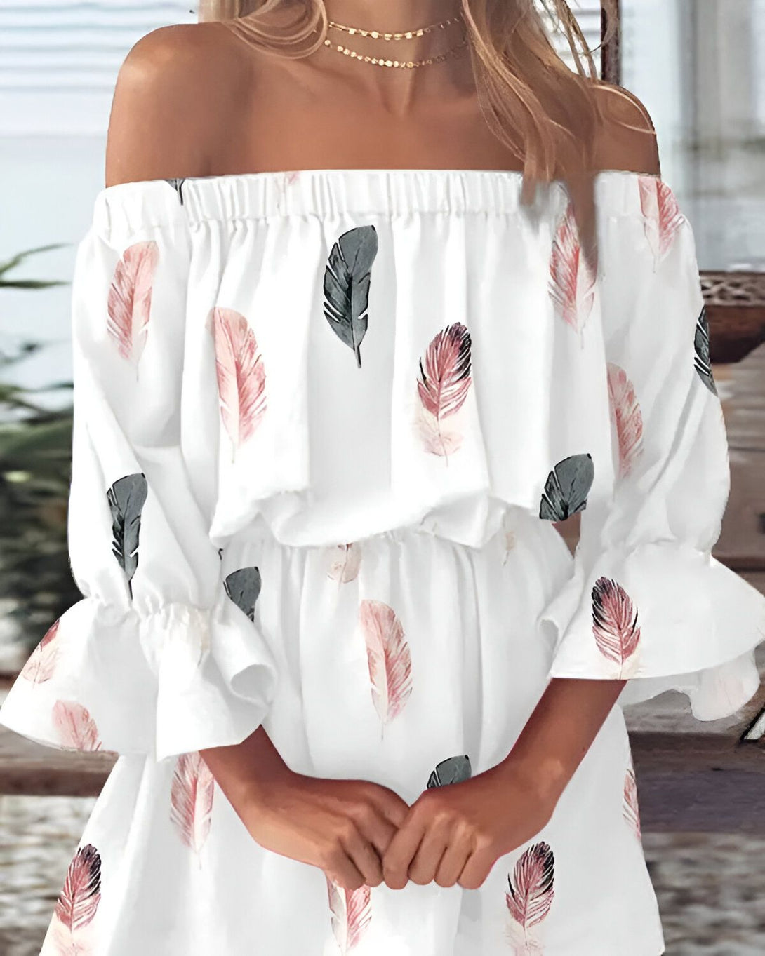 Une jeune femme porte une robe blanche à épaules dénudées. La robe est imprimée de plumes roses et grises. La femme a les cheveux longs et blonds. Elle porte un collier et des boucles d'oreilles. Elle est debout dans une pièce, adossée à une table en bois.