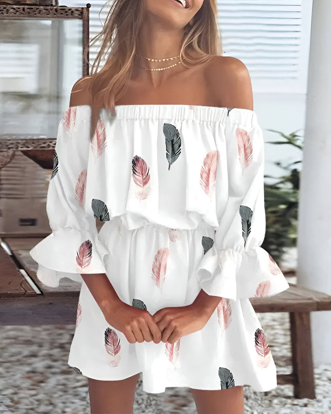Une jeune femme porte une robe blanche à épaules dénudées. La robe est imprimée de plumes roses et grises. La femme a les cheveux longs et blonds et elle sourit. Elle porte un collier et des bracelets.
