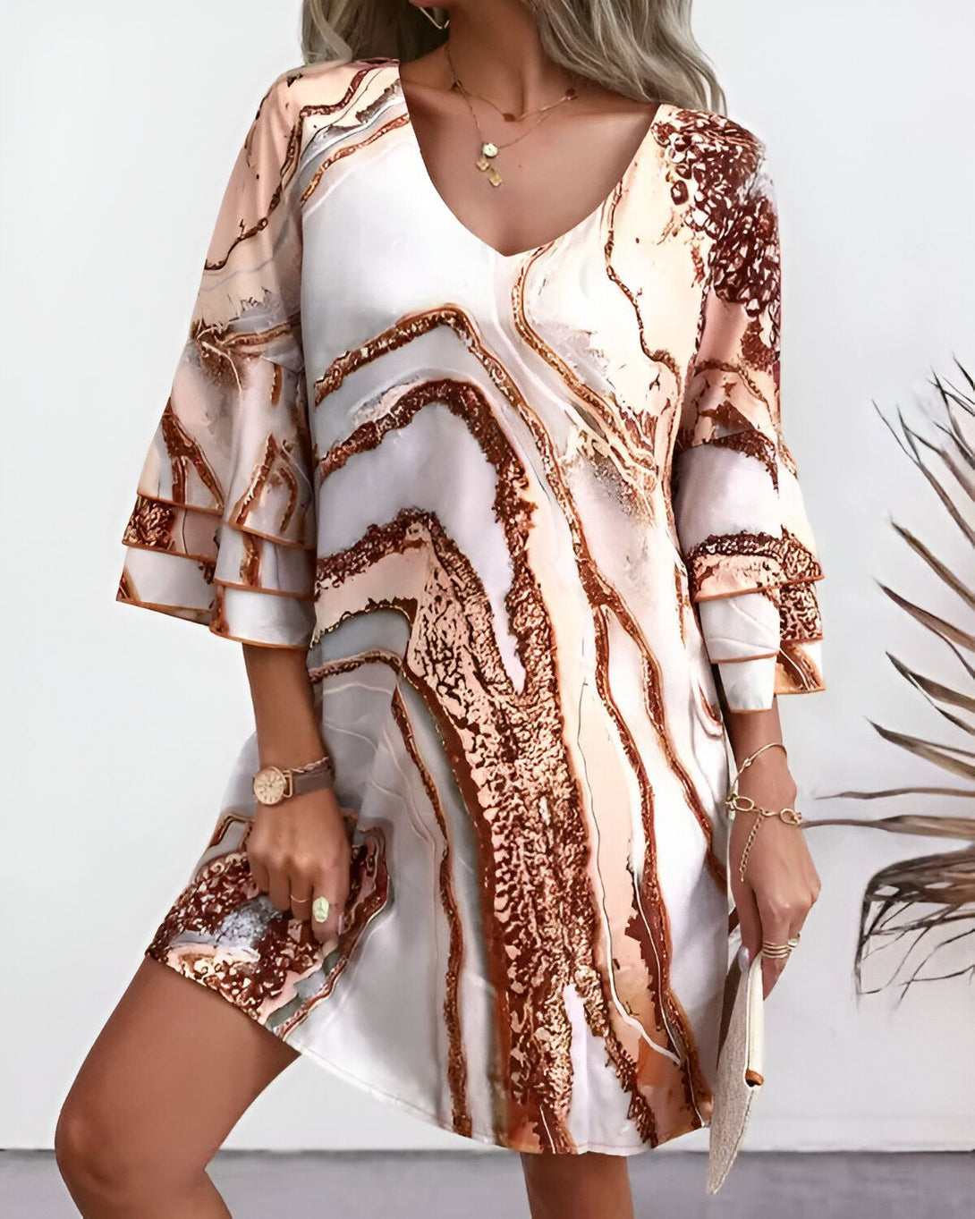 Une femme porte une robe mi-longue à manches 3/4. La robe est blanche avec des motifs marbrés marron et or. La femme est blonde et porte un collier, une montre et des bracelets. Elle porte également un sac à main marron.