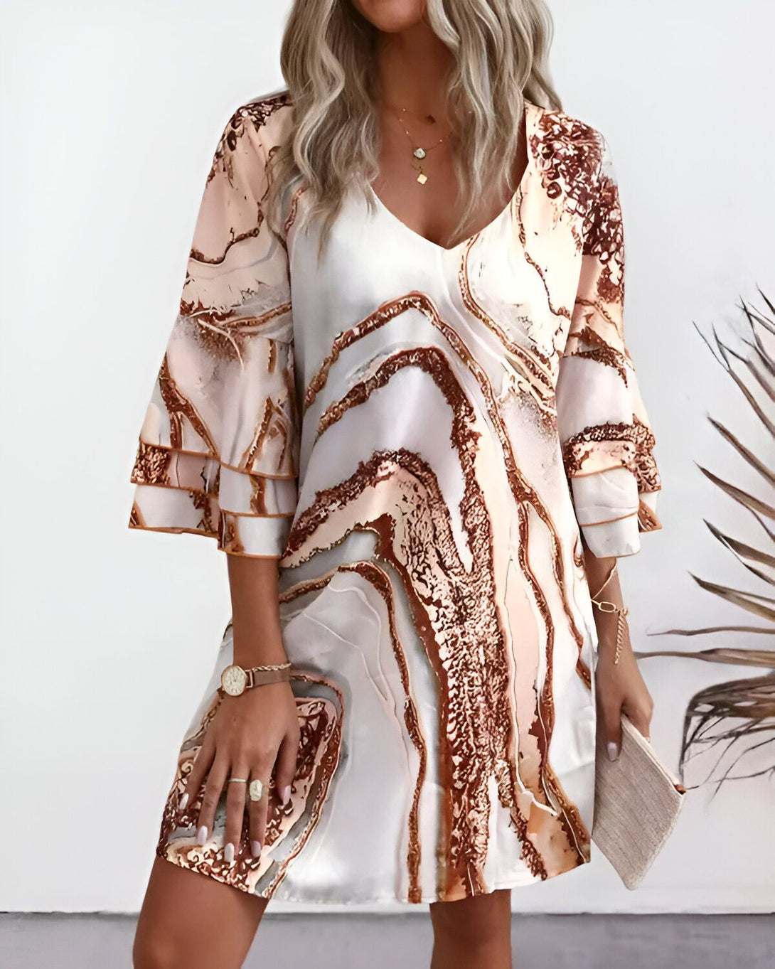 Une femme porte une robe blanche et marron. La robe est courte et a des manches bouffantes. La femme est blonde et a les cheveux longs. Elle porte un collier et un bracelet. Elle est debout dans une pièce et il y a un palmier derrière elle.