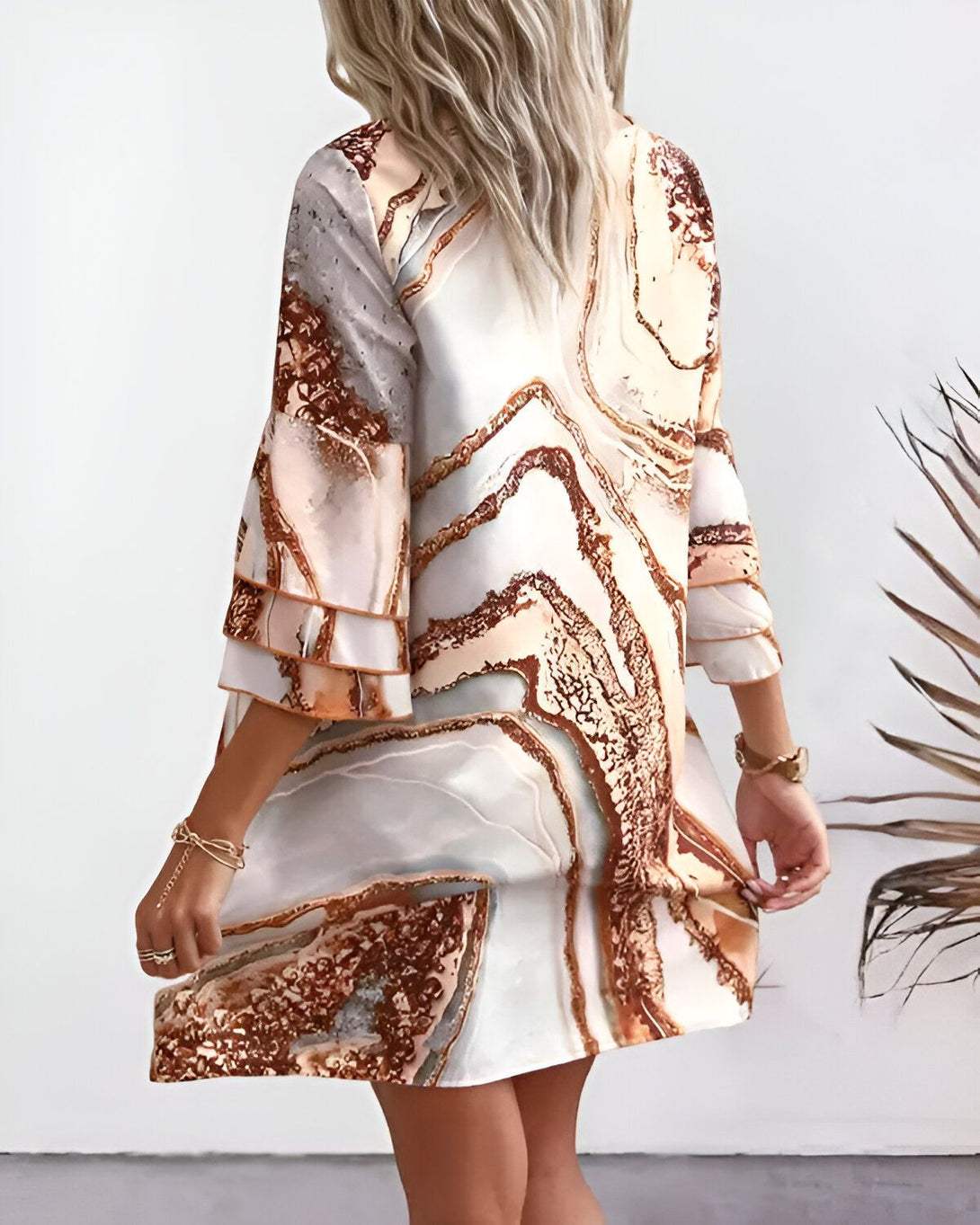 Une femme porte une robe mi-longue à manches 3/4. La robe est cintrée à la taille et évasée en bas. Elle est de couleur marron et blanche avec un motif marbré. La femme est blonde et porte des talons hauts marron. Elle se tient debout, de dos, face à l'appareil photo.