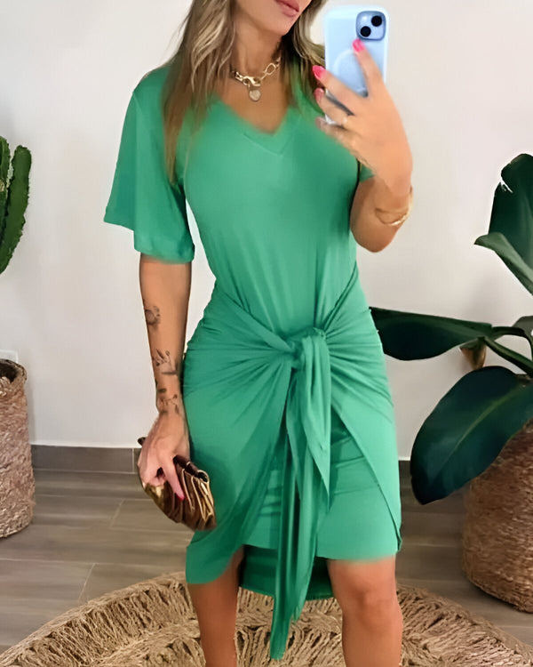 Une femme porte une robe verte courte à manches courtes. Elle est ceinturée à la taille et nouée sur le côté. Elle porte un collier et un bracelet. Elle tient un téléphone portable dans sa main droite et un sac à main doré dans sa main gauche. Elle est debout sur un tapis rond en osier, dans un salon, et prend un selfie.