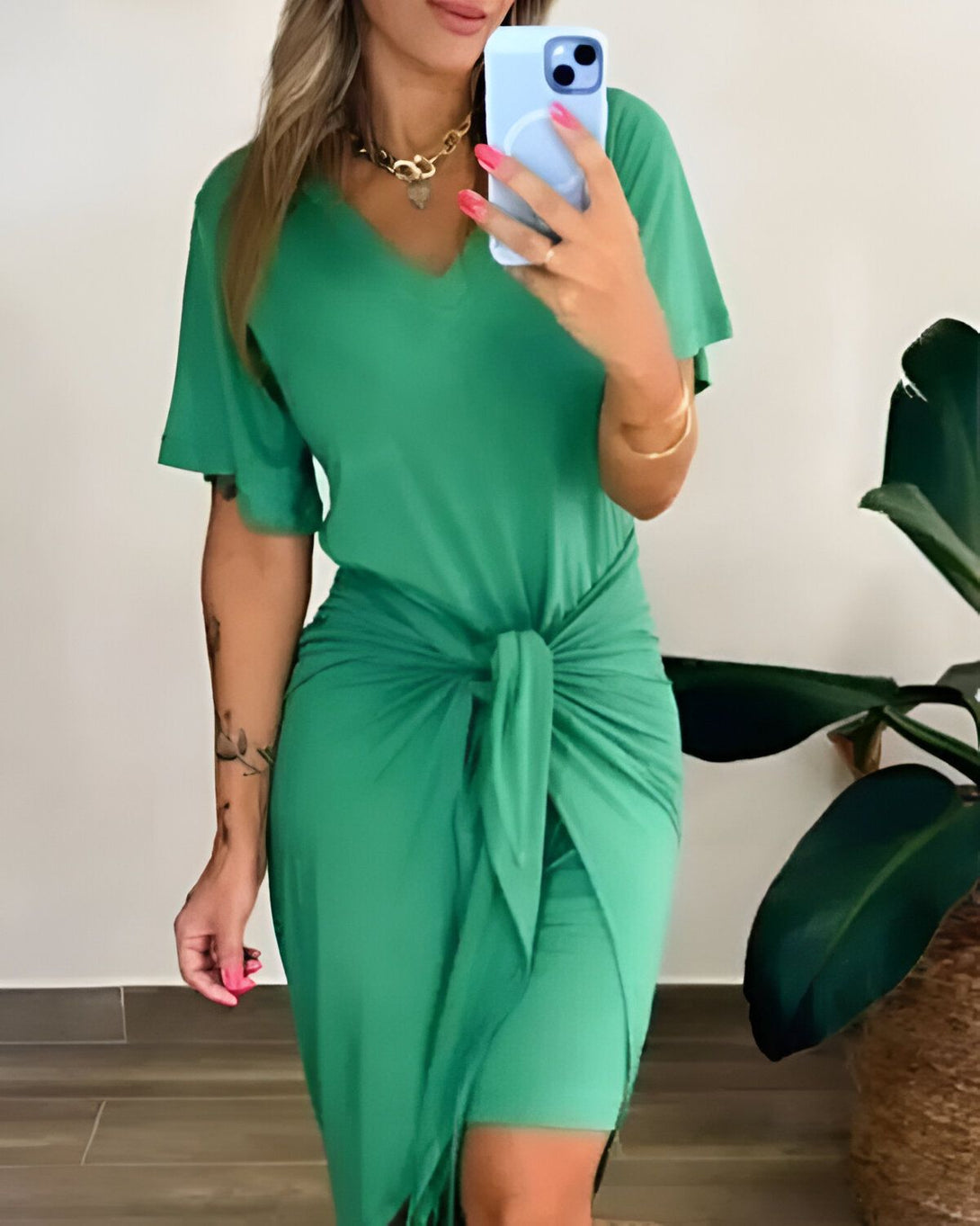 Une femme porte une robe verte courte avec un décolleté en V et des manches courtes. Elle est ceinturée à la taille et nouée sur le côté. Elle porte un collier et des bracelets.