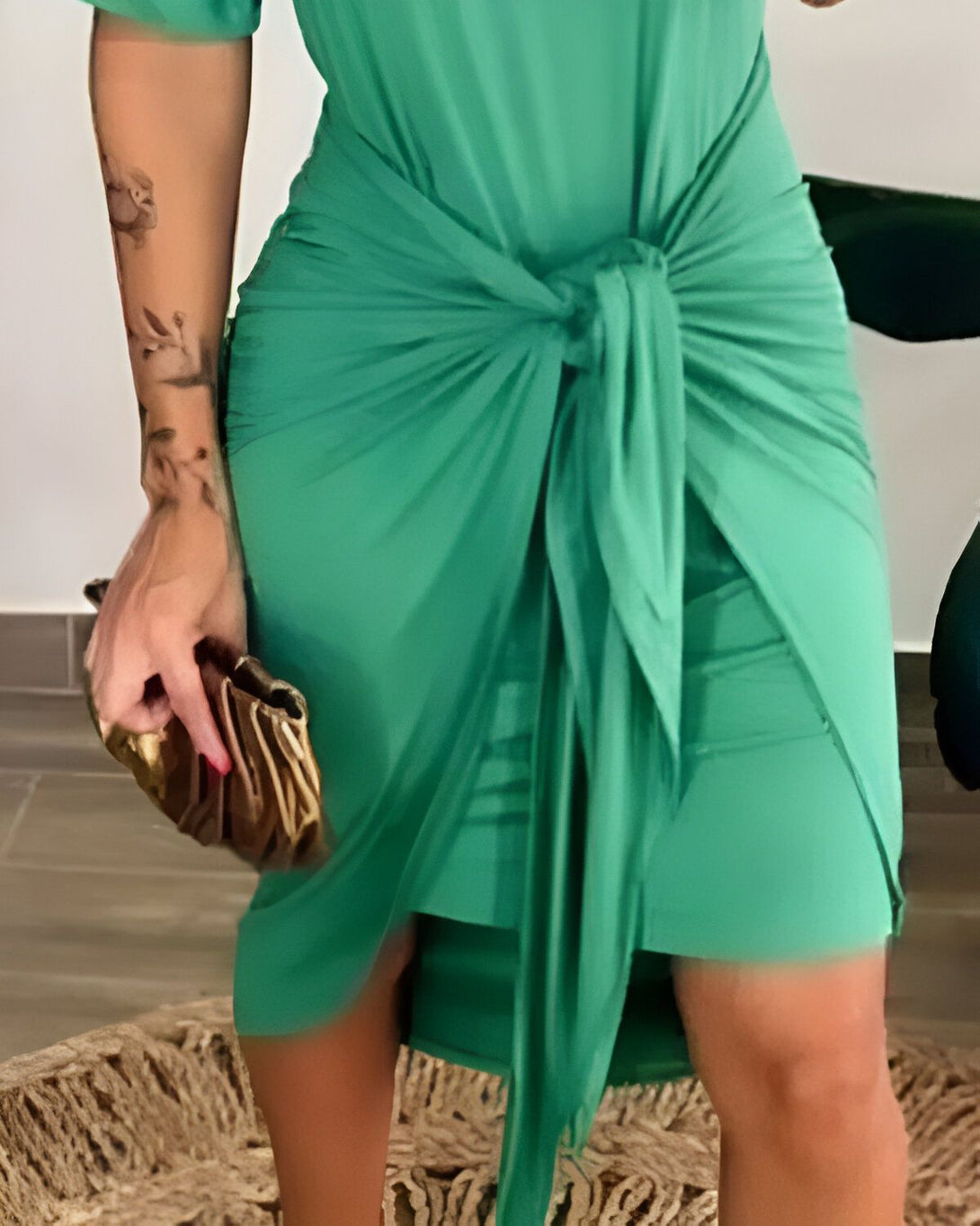 Une femme porte une robe verte ajustée nouée sur le côté. Elle porte également des talons hauts dorés et tient un sac à main doré.