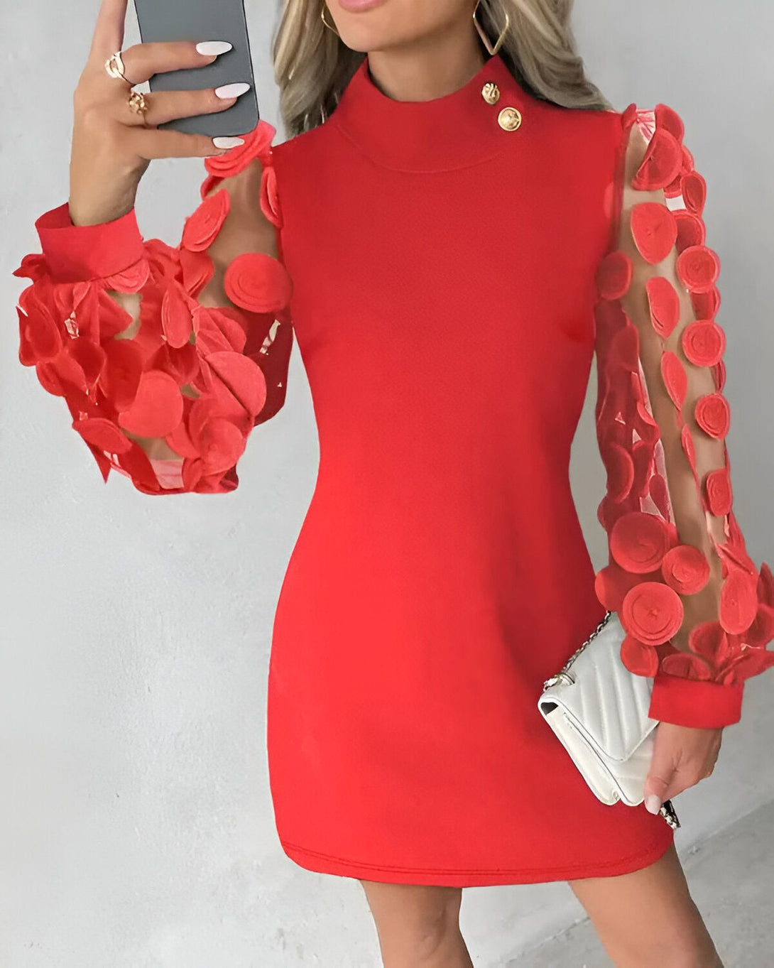 Une femme porte une robe rouge moulante à manches longues. Les manches sont en résille et transparentes. La robe est courte et s'arrête au-dessus du genou. La femme est blonde et porte un maquillage léger. Elle a les ongles manucurés et porte une bague à la main droite. Elle tient un téléphone portable dans la main gauche.