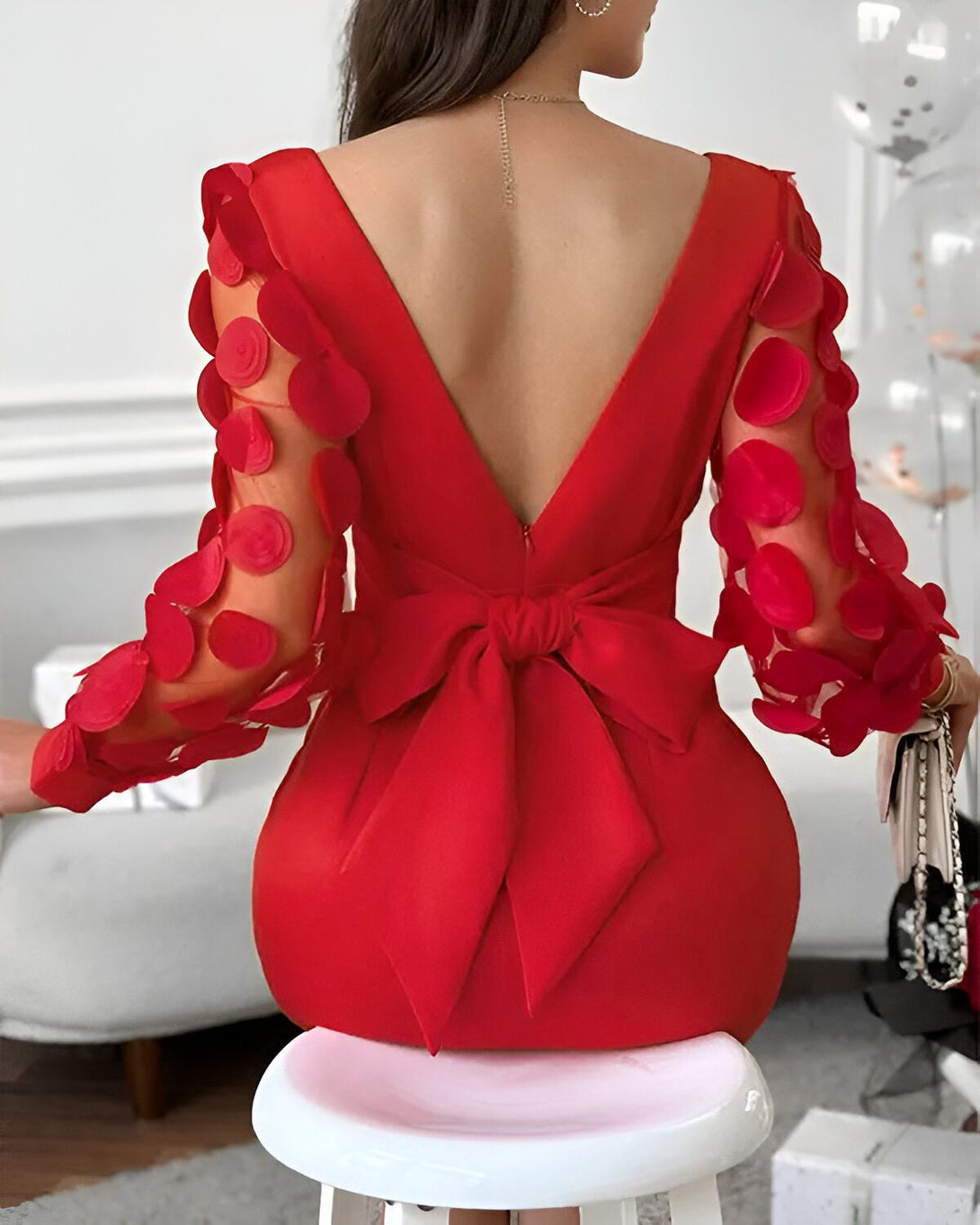 Une femme portant une robe rouge moulante avec un grand nœud rouge dans le dos. La robe est courte et a des manches longues. La femme est assise sur un tabouret et porte des talons hauts rouges.
