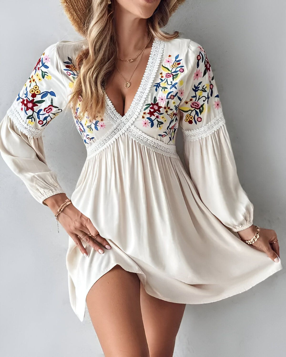 Une jeune femme porte une robe blanche courte à manches longues. La robe est décolletée en V et brodée de fleurs colorées. La femme est blonde et porte un chapeau de paille. Elle est souriante et regarde l'objectif.
