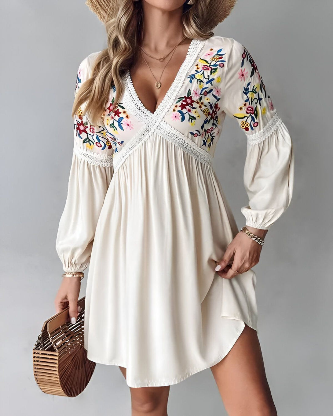 Une jeune femme porte une robe blanche courte à manches longues. La robe est brodée de fleurs colorées. Elle porte un chapeau de paille et tient un sac à main en osier.