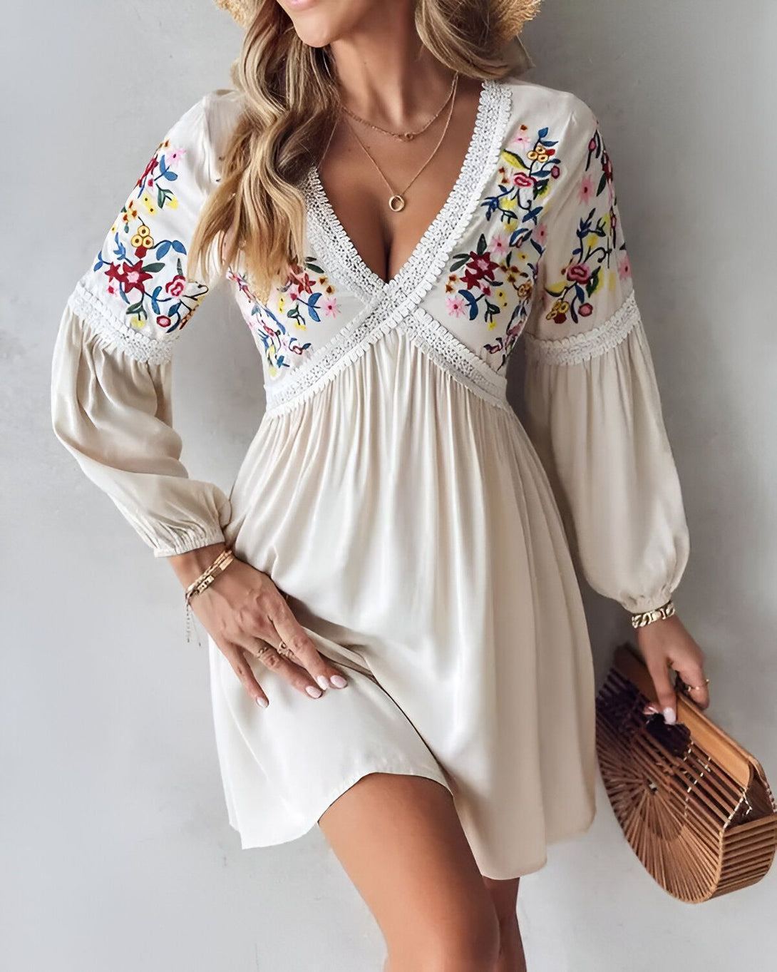 Une jeune femme porte une robe blanche brodée de fleurs. La robe est courte et a des manches longues. La femme est blonde et porte un chapeau de paille. Elle est souriante et tient un sac à main marron.
