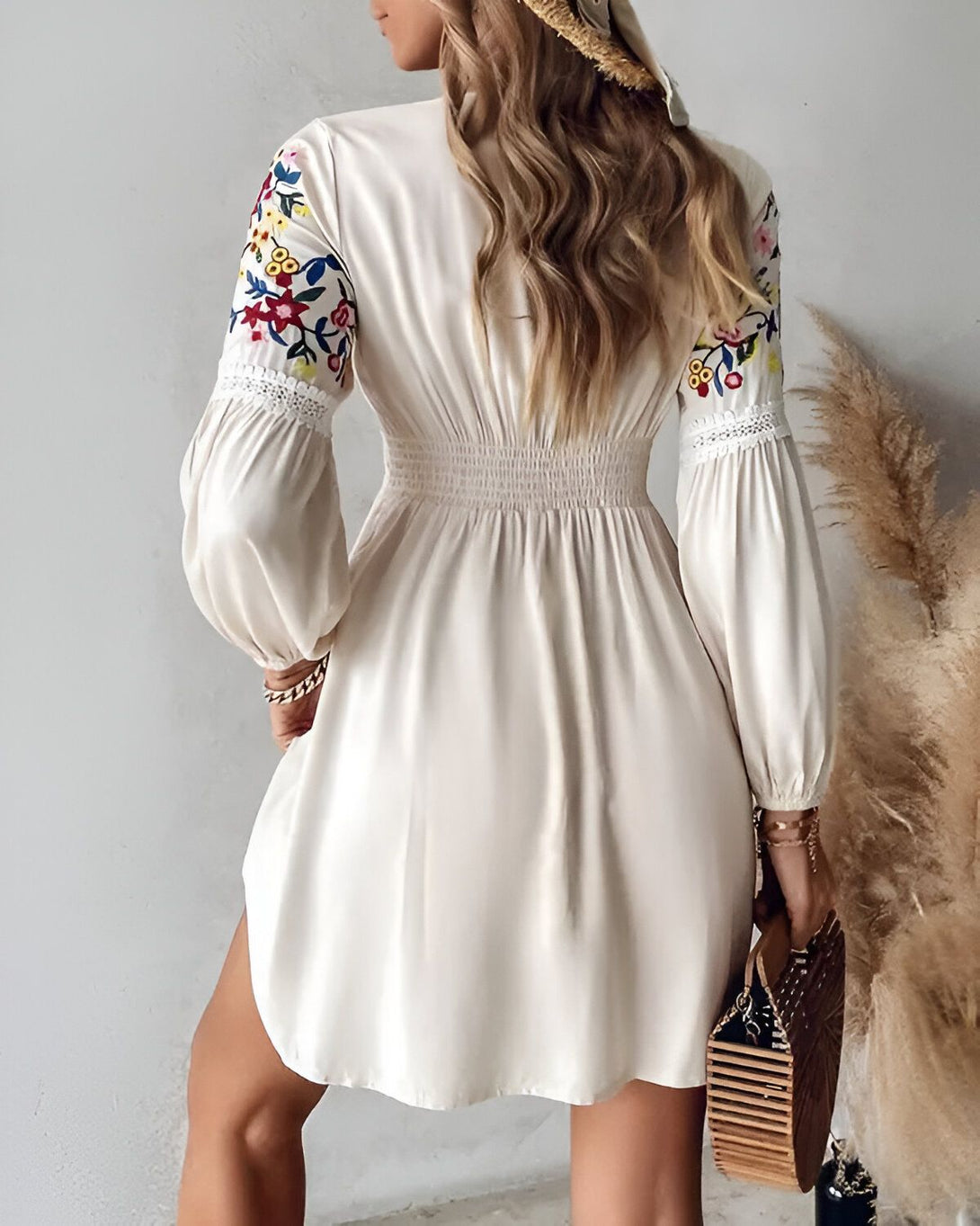 Une femme porte une robe blanche courte à manches longues. La robe est cintrée à la taille et possède un décolleté en V. Elle est également brodée de fleurs colorées. La femme porte un chapeau de paille et tient un sac à main en osier.