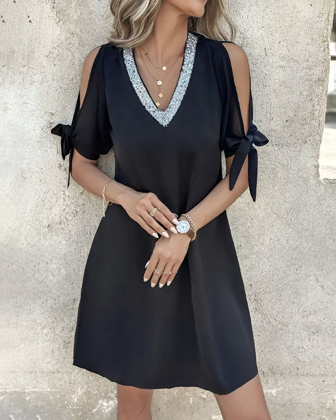 Une jeune femme porte une robe noire avec des sequins argentés au niveau du col. Elle a les cheveux blonds et les yeux bleus. Elle est souriante et regarde l'objectif.