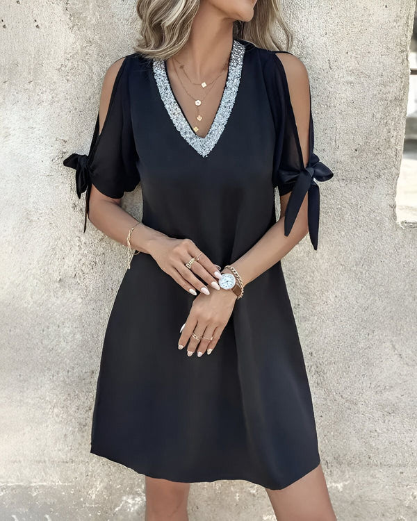 Une jeune femme porte une robe noire avec des sequins argentés au niveau du col. Elle a les cheveux blonds et les yeux bleus. Elle est souriante et regarde l'objectif.