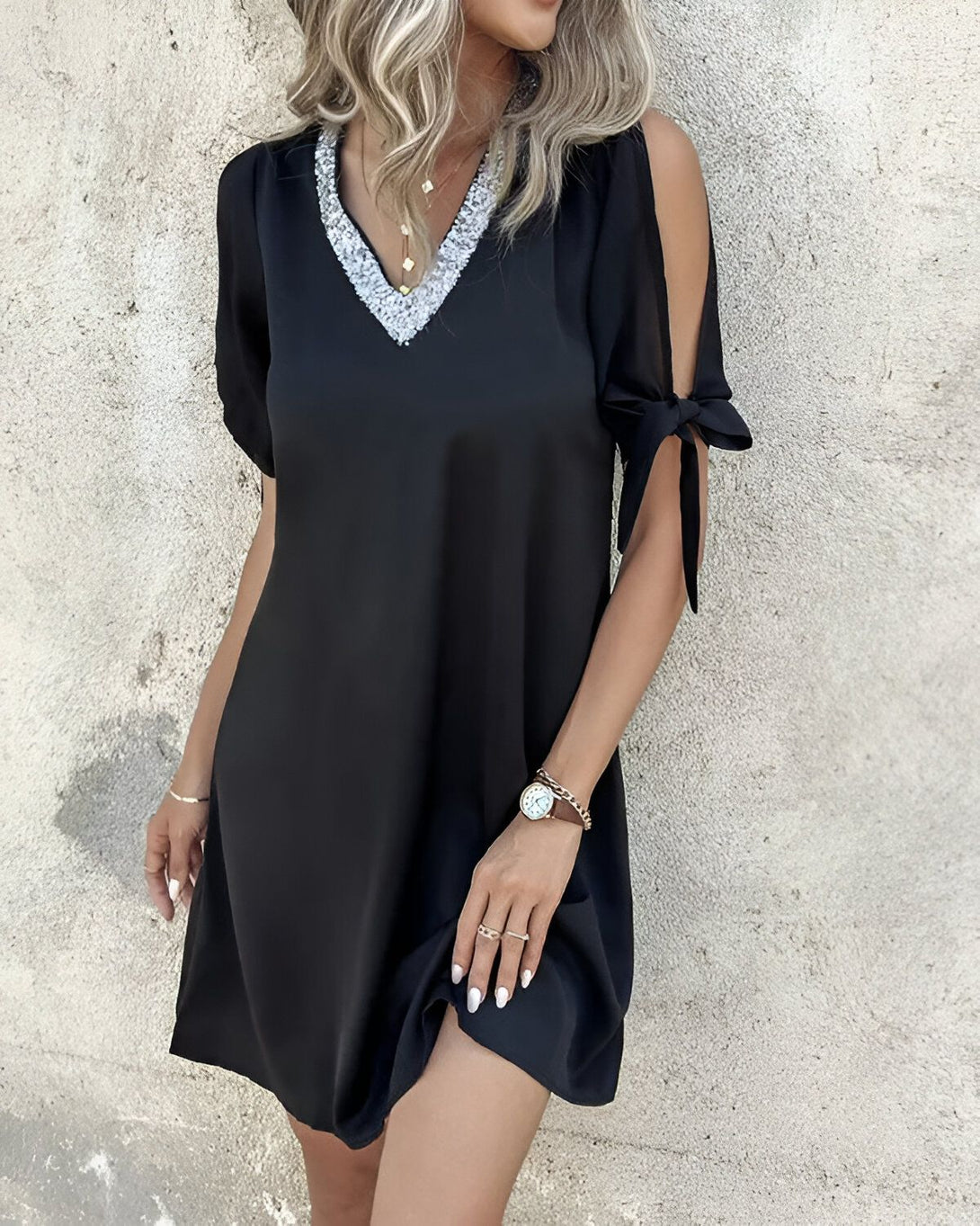 Une femme porte une robe noire avec des sequins argentés au niveau du col. La robe est courte et les manches sont nouées au niveau des épaules. La femme porte également un collier et une montre.