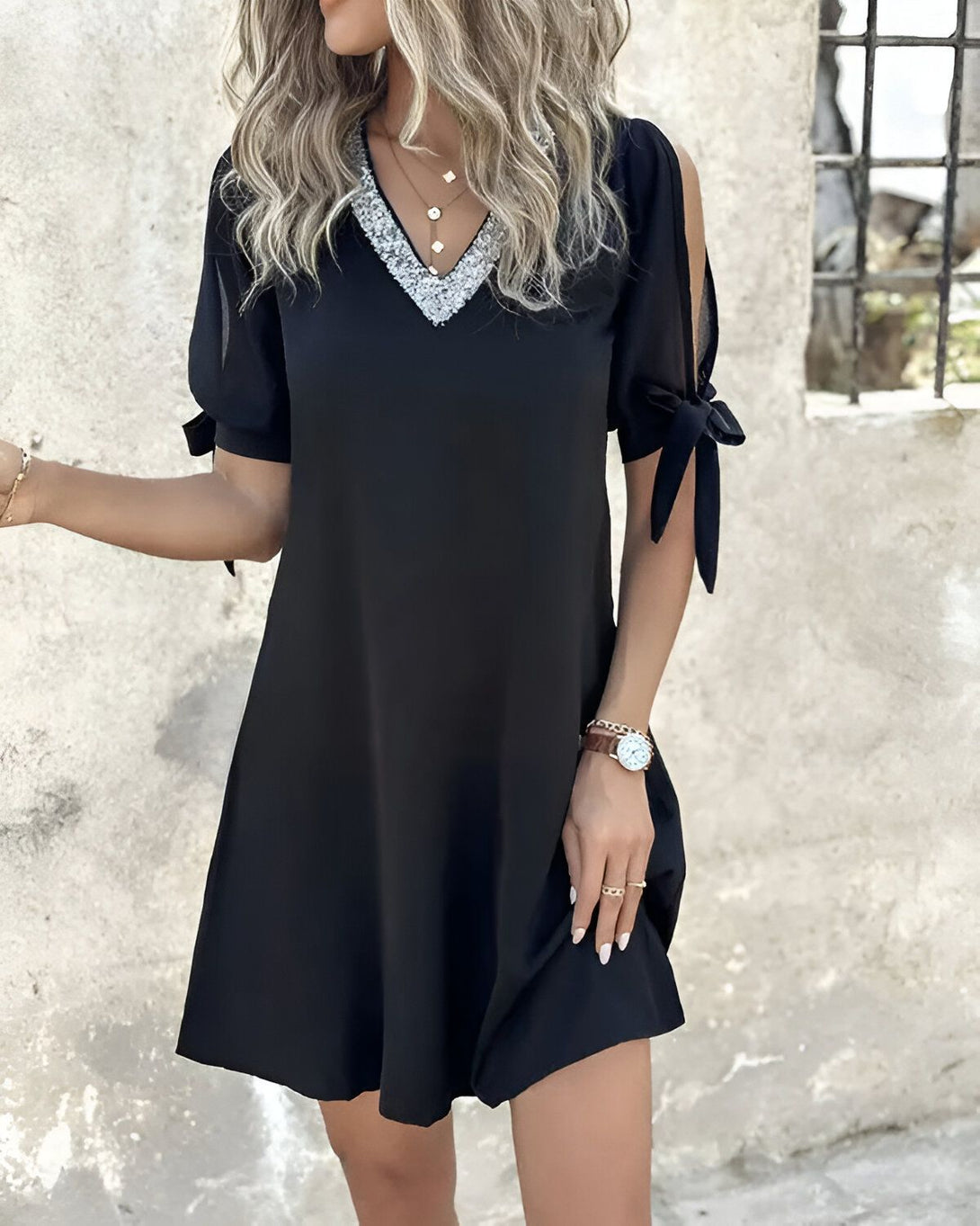 Une jeune femme portant une robe noire avec des manches bouffantes et un col en V. La robe est courte et évasée. La femme est blonde et a les cheveux longs. Elle porte un collier et un bracelet. Elle est debout devant un mur de pierre.