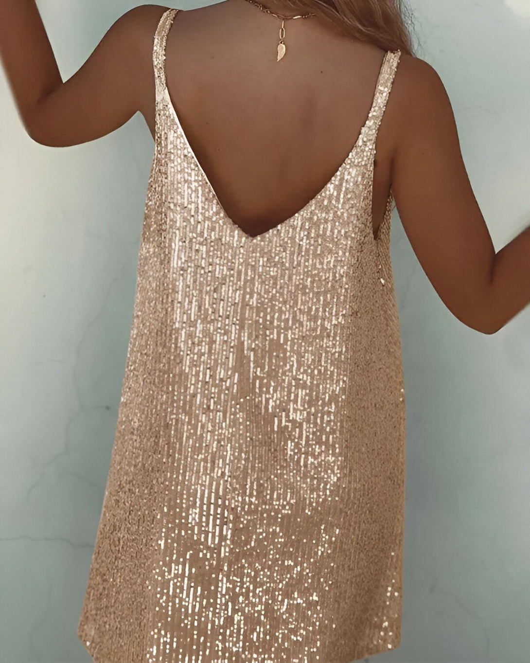 Une femme porte une robe dos nu à sequins dorés. La robe est courte et a des bretelles fines. La femme est vue de dos et ses cheveux sont longs et blonds. Elle porte un collier avec un pendentif en forme de feuille.
