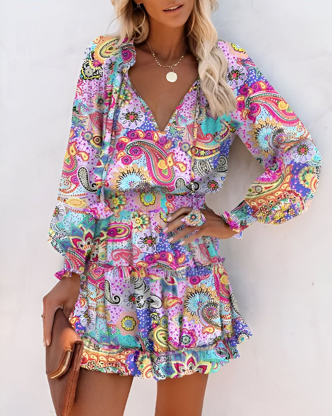 Une femme porte une robe courte à manches longues et à volants. La robe est rose et violette, avec un motif cachemire. La femme est blonde et porte un collier. Elle a les cheveux lâchés et un maquillage léger. Elle est souriante et regarde l'appareil photo.