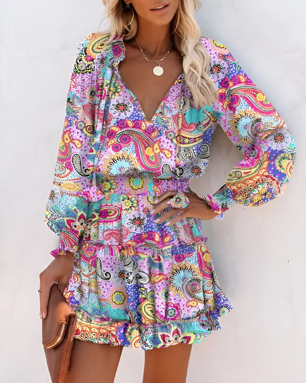 Une femme porte une robe courte à manches longues et à volants. La robe est rose et violette, avec un motif cachemire. La femme est blonde et porte un collier. Elle a les cheveux lâchés et un maquillage léger. Elle est souriante et regarde l'appareil photo.