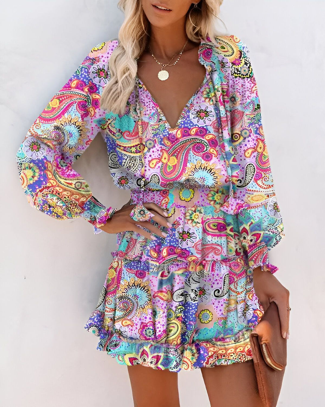 Une femme porte une robe colorée à motifs cachemire. La robe est courte et évasée, avec un décolleté en V et des manches longues. La femme est blonde et porte un collier. Elle est debout devant un mur blanc.