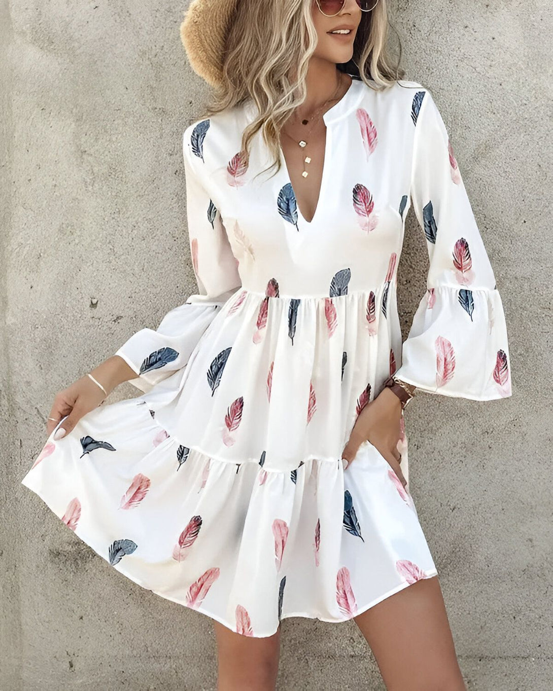 Une jeune femme porte une robe blanche à motifs de plumes colorées. Elle a un chapeau de paille sur la tête et des lunettes de soleil. Elle est souriante et a l'air heureuse.