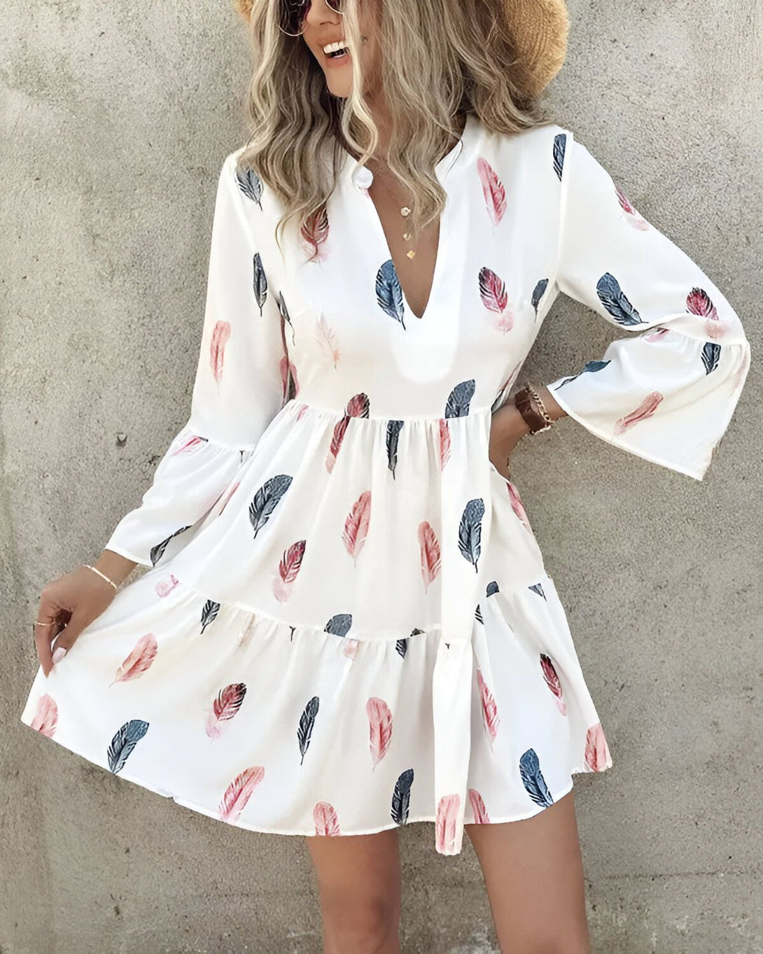 Une femme porte une robe blanche à motifs de plumes colorées. La robe est courte et évasée, avec des manches longues et un col en V. La femme est souriante et porte un chapeau de paille.