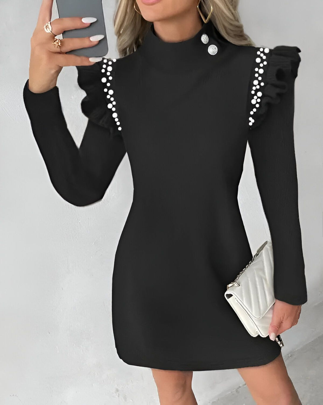 Une femme porte une robe noire avec des perles sur les épaules. La robe est moulante et a un col montant. La femme a les cheveux blonds et porte un maquillage léger. Elle tient un téléphone portable dans sa main droite et un sac à main blanc dans sa main gauche.
