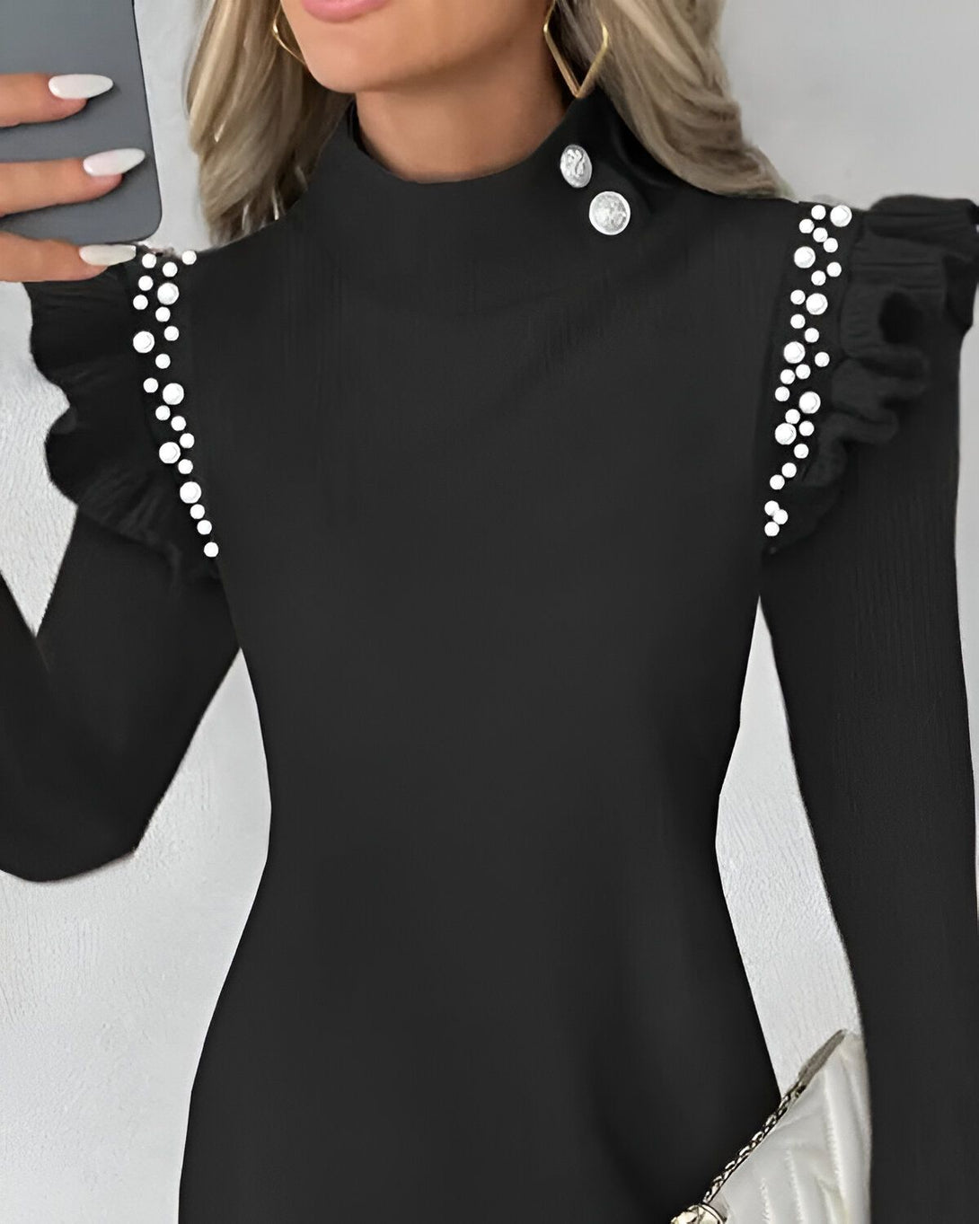 Une femme porte une robe noire à manches longues avec des perles sur les épaules. La robe est ajustée et a un col montant. La femme a les cheveux blonds et porte un maquillage léger. Elle tient un téléphone portable dans sa main droite.
