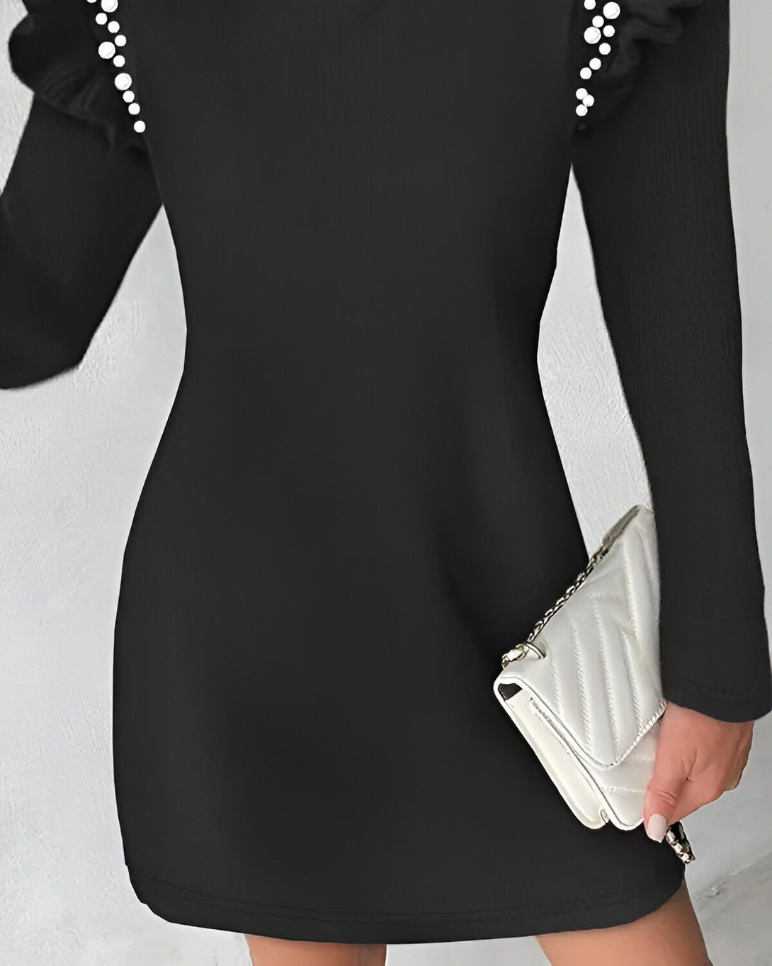 Une femme porte une robe noire moulante avec des perles sur les épaules et des manches longues. Elle porte également un sac à main blanc.