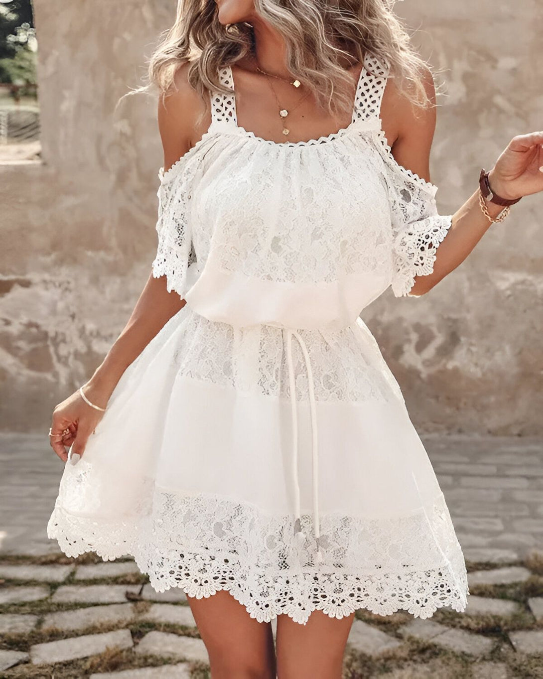 Une jeune femme portant une robe blanche en dentelle. La robe est courte et évasée, avec un décolleté en V et des bretelles fines. La femme a les cheveux blonds et elle est souriante. Elle se tient debout dans une rue pavée, avec un mur de pierre en arrière-plan.