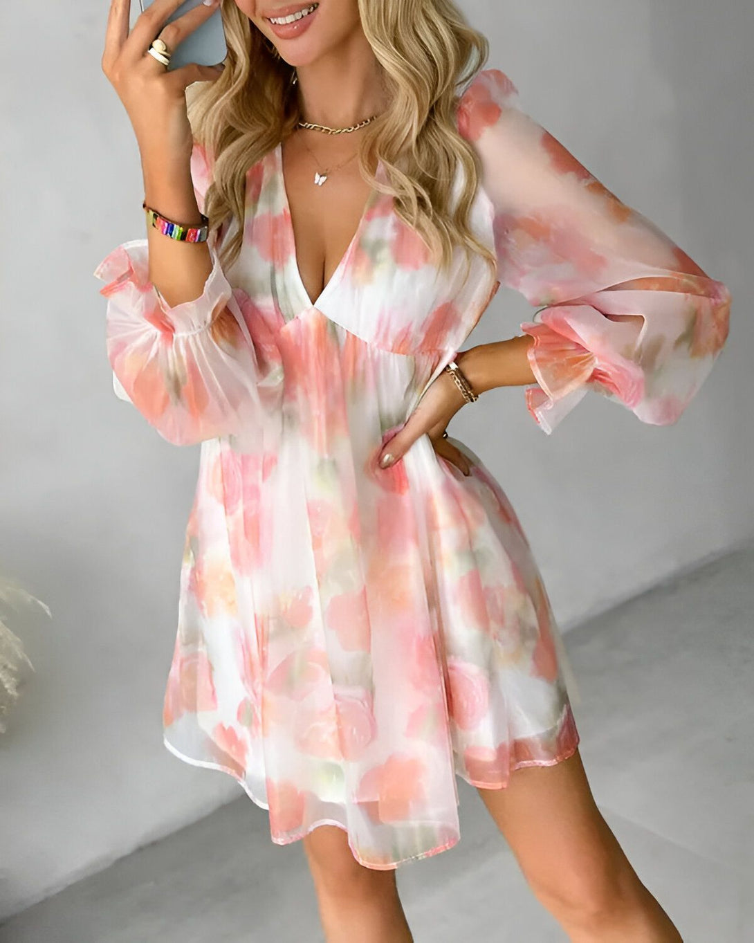 Une jeune femme blonde porte une robe courte en mousseline de soie blanche à motifs floraux roses. La robe a un décolleté en V et des manches longues bouffantes. La femme est souriante et porte des bracelets en perles et en or.
