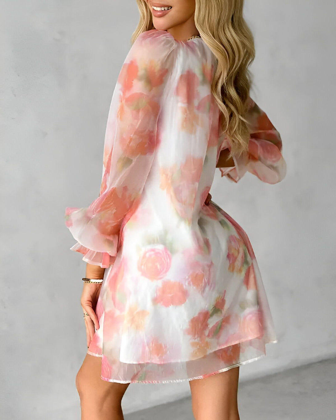Une jeune femme portant une robe blanche transparente à motifs floraux roses. La robe a des manches bouffantes et un ourlet volanté. La femme est blonde et a les cheveux longs et ondulés. Elle porte un bracelet doré au poignet gauche.