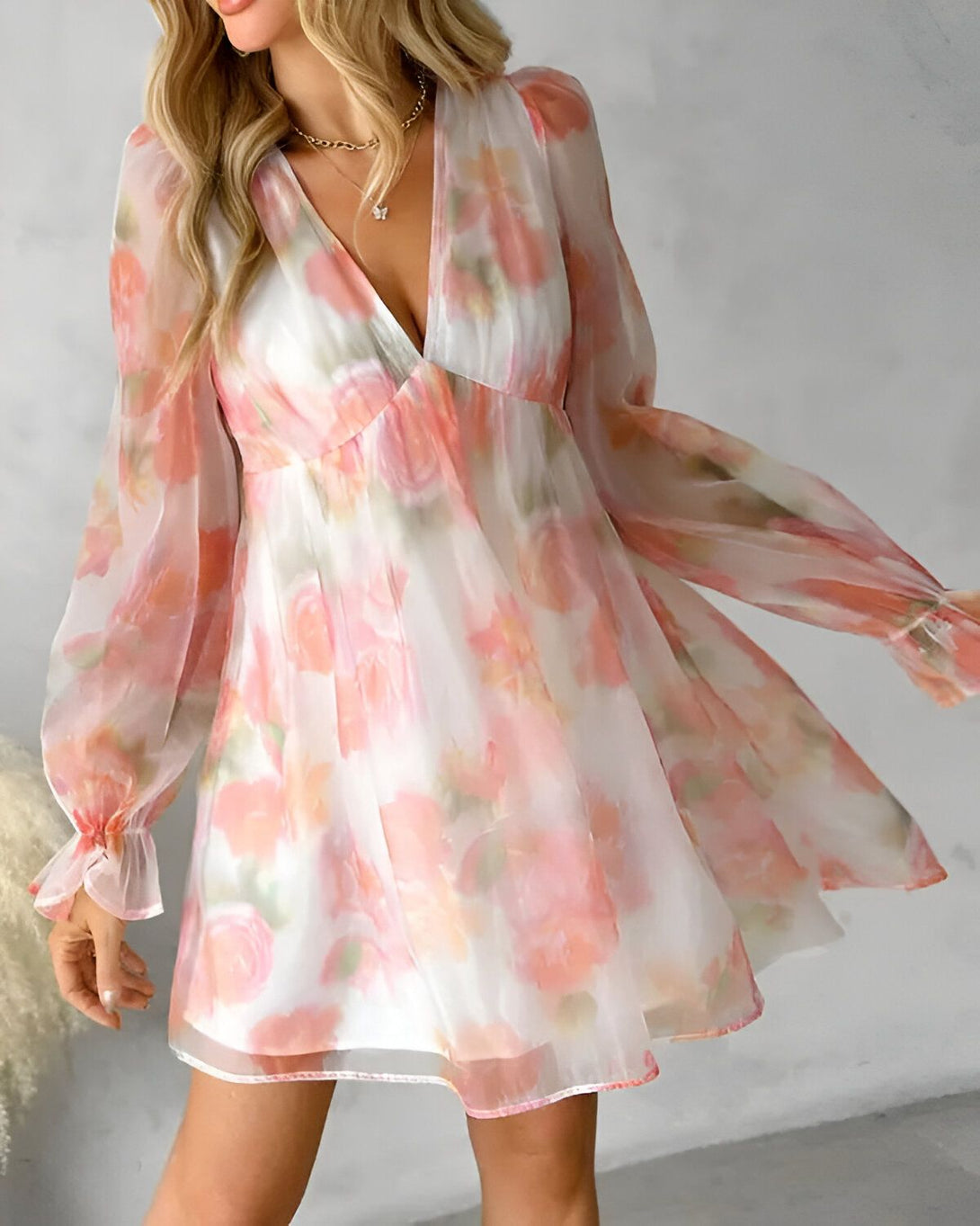 Une jeune femme porte une robe courte en mousseline de couleur rose et blanche. La robe est transparente et l'on peut voir ses jambes à travers. Elle a les cheveux longs et blonds et porte un collier doré. Elle est debout dans un studio et sourit à la caméra.