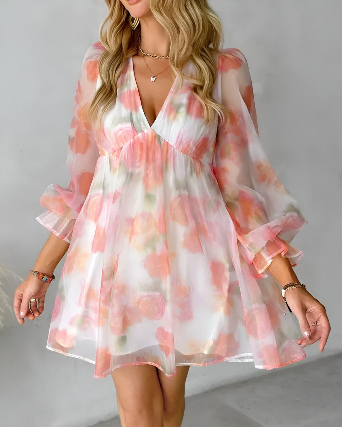 Une jeune femme porte une robe courte en mousseline de couleur blanche à motifs de fleurs roses. La robe est décolletée en V et possède des manches longues et bouffantes.