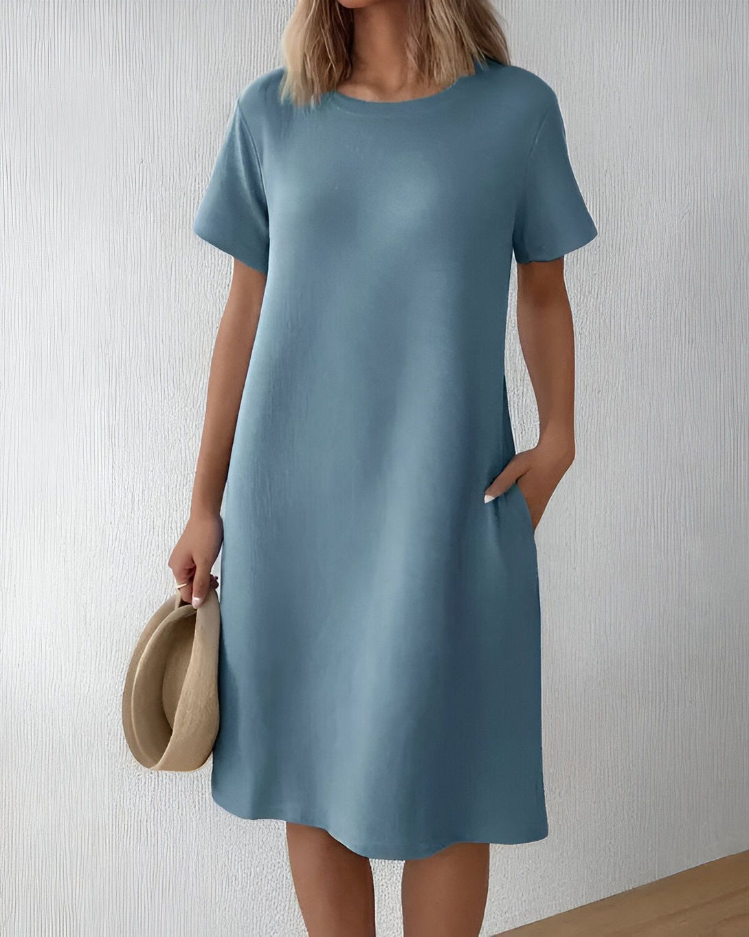 Une jeune femme porte une robe bleu clair. La robe est courte et a des manches courtes. La femme a les cheveux blonds et elle porte un chapeau de paille. Elle est debout devant un mur blanc.