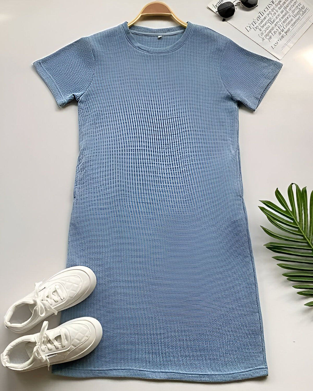Une robe bleu clair à manches courtes avec des baskets blanches et des lunettes de soleil sur un fond blanc.
