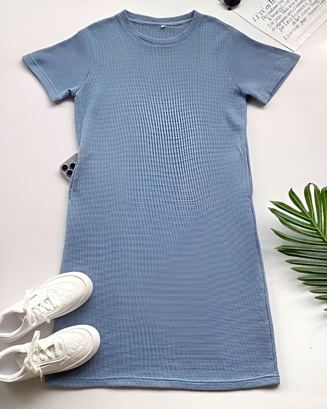 Une robe bleu clair à manches courtes avec des baskets blanches et des lunettes de soleil sur un fond blanc.