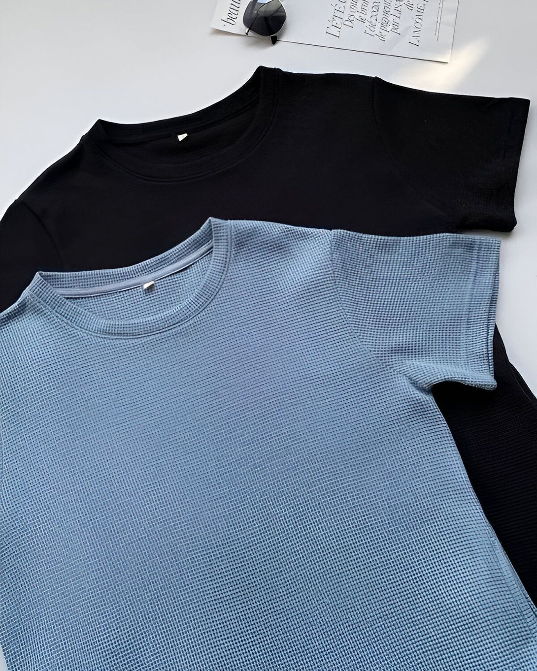 Deux t-shirts en coton, un bleu et un noir, sont posés à plat sur un fond blanc.