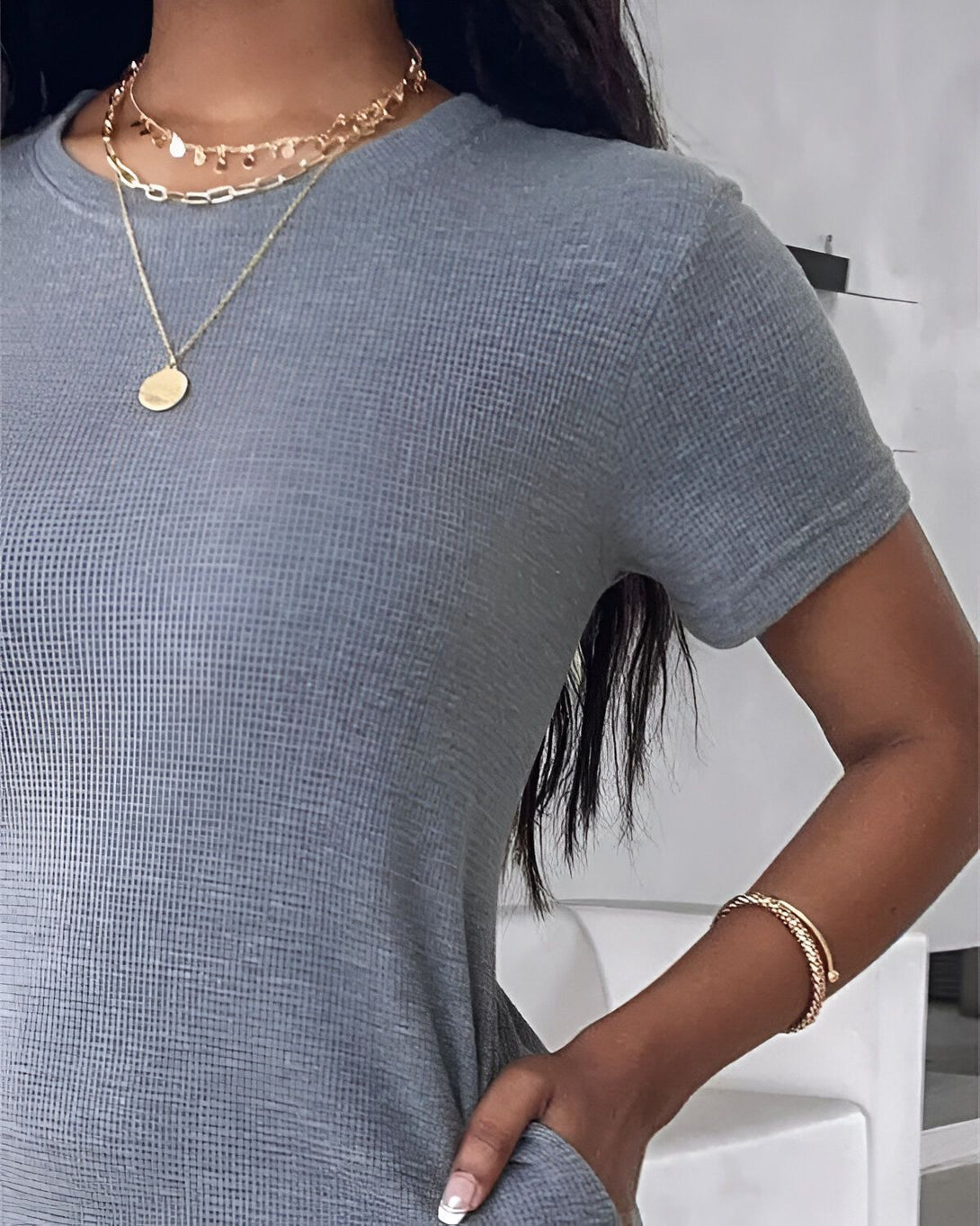 Une femme porte un t-shirt gris avec un collier et un bracelet en or.