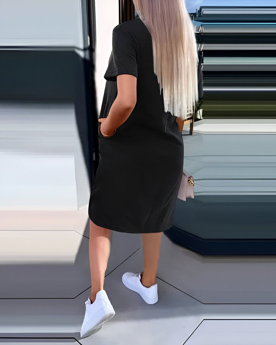 Une femme blonde porte une robe noire décontractée avec des baskets blanches. Elle a les mains dans les poches et porte un sac à main rose.