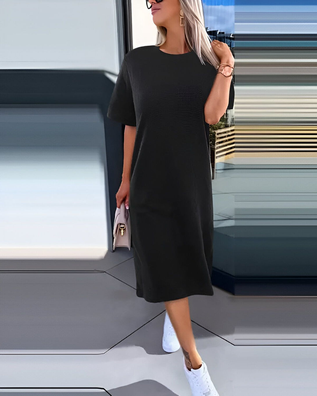 Une femme porte une robe noire mi-longue près du corps à manches courtes. Elle est assortie d'une paire de baskets blanches et d'un sac à main beige.