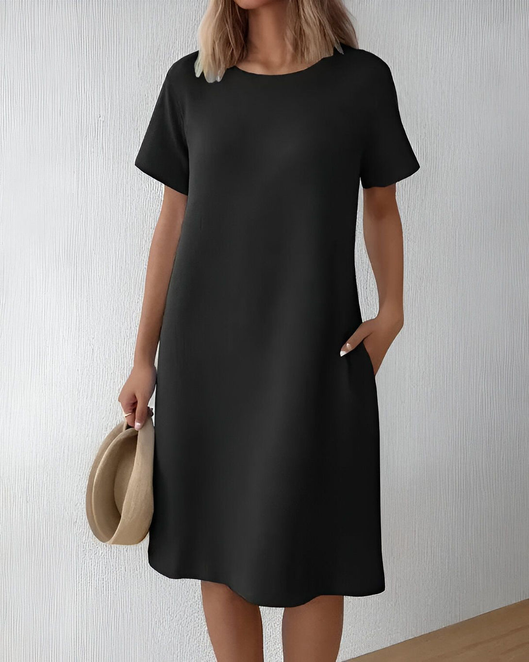 Une jeune femme porte une robe noire simple avec des manches courtes et des poches. Elle porte également un chapeau de paille marron et tient un autre chapeau de paille marron à la main.