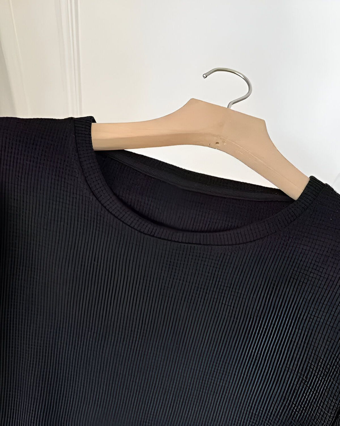 Une image d'un cintre en bois avec un t-shirt noir texturé accroché dessus. Le t-shirt est à col rond et a des manches courtes.