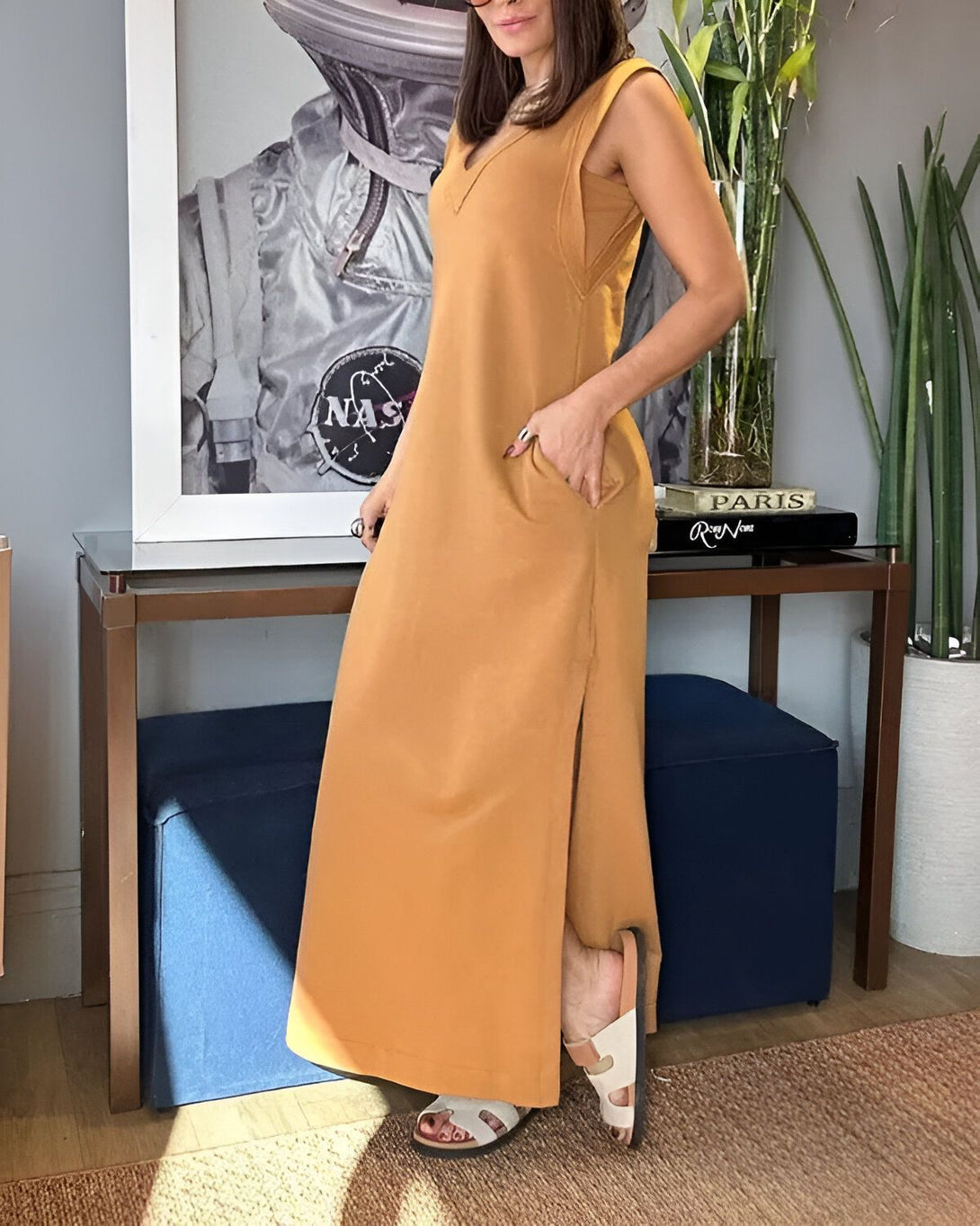 Une femme porte une longue robe marron sans manches avec des sandales marron clair. Elle a les cheveux longs et bruns et porte du maquillage. Elle se tient debout dans un salon, avec une main dans la poche et l'autre tenant le haut de la robe.