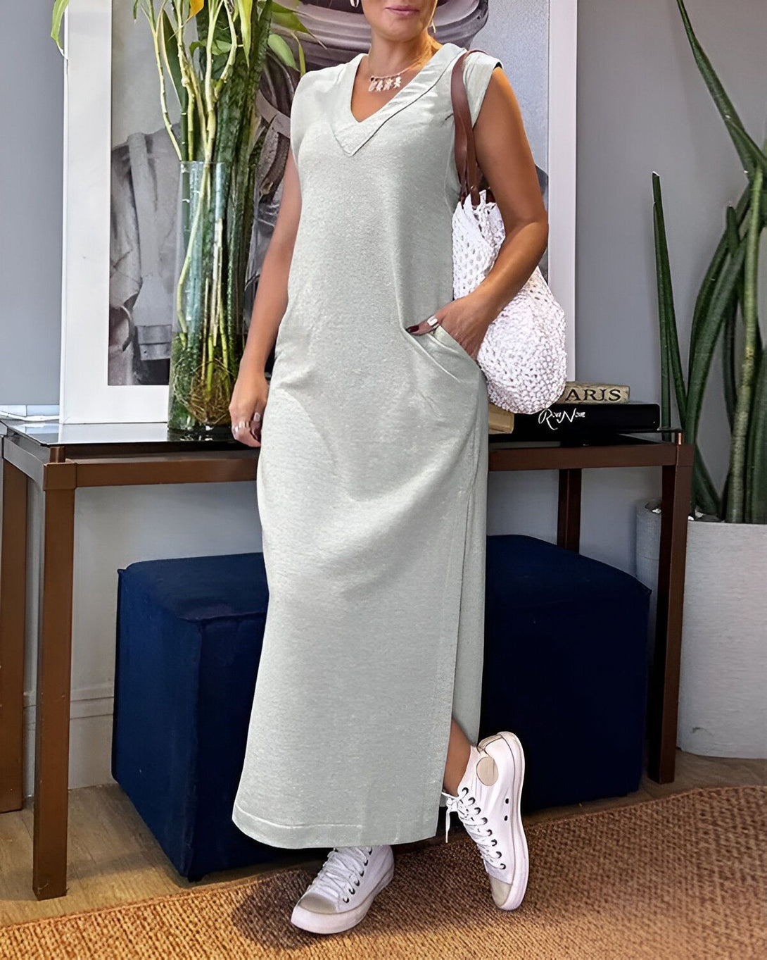 Une femme porte une robe longue vert clair sans manches avec des baskets blanches et un sac à main en osier blanc. Elle est debout dans un salon, il y a des plantes vertes et des cadres accrochés au mur.
